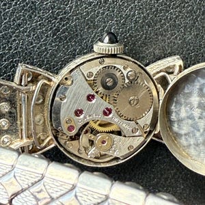 May include: Close-up of a vintage silver wristwatch with an open face, revealing the intricate mechanical movement. The watch features a black gem on the crown and a silver band with a geometric design. The watch face is marked "17 JEWELS SWISS MADE".