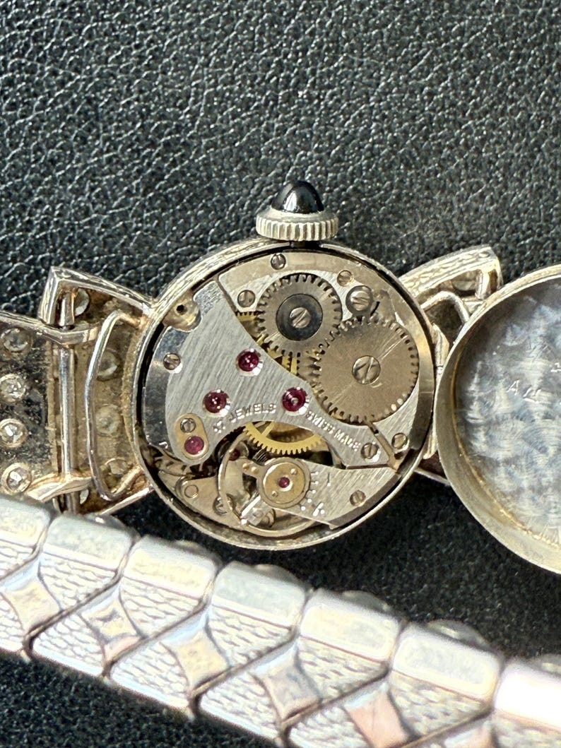 May include: Close-up of a vintage silver wristwatch with an open face, revealing the intricate mechanical movement. The watch features a black gem on the crown and a silver band with a geometric design. The watch face is marked "17 JEWELS SWISS MADE".