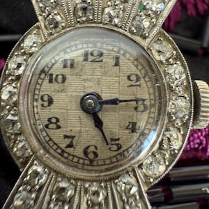 May include: Close-up of a vintage silver wristwatch with a round face. The watch features a diamond-encrusted bezel and band, with a textured, off-white watch face. Black numerals mark the hours. The hands point to approximately 3:15.