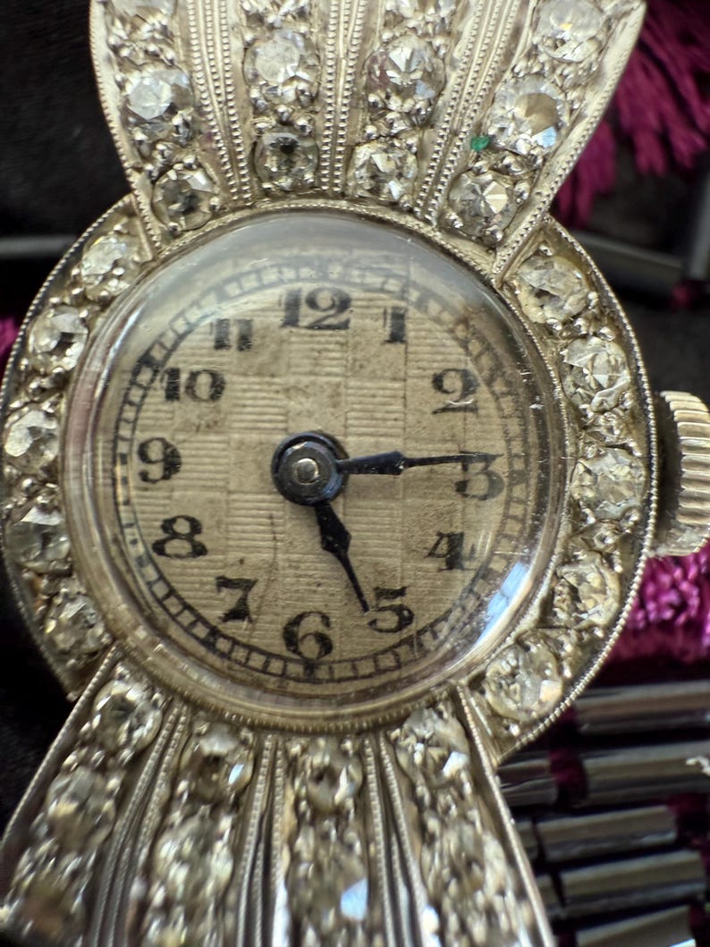 May include: Close-up of a vintage silver wristwatch with a round face. The watch features a diamond-encrusted bezel and band, with a textured, off-white watch face. Black numerals mark the hours. The hands point to approximately 3:15.