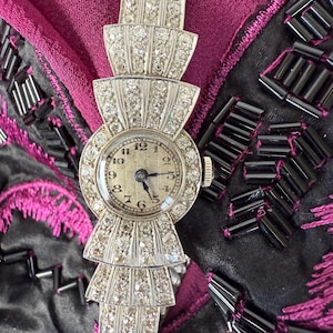 May include: A vintage silver wristwatch with a round face and a diamond-encrusted bezel. The watch band features a tiered, fan-like design, also adorned with diamonds. The watch hands point to 10:09. The watch is set against a backdrop of dark fabric.