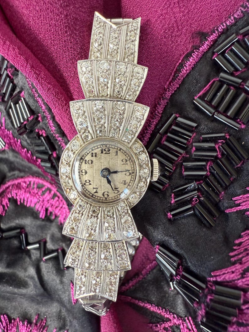 May include: A vintage silver wristwatch with a round face and a diamond-encrusted bezel. The watch band features a tiered, fan-like design, also adorned with diamonds. The watch hands point to 10:09. The watch is set against a backdrop of dark fabric.