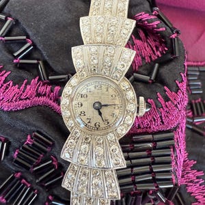 May include: A vintage silver-toned wristwatch with a round face and a decorative band. The watch face has black numerals and hands. The band features multiple tiers of sparkling, clear gemstones. The watch is set against a black and magenta background.