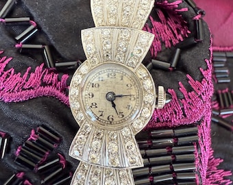 Könnte beinhalten: Eine Vintage-Armbanduhr in Silberoptik mit rundem Zifferblatt und dekorativem Armband. Das Zifferblatt hat schwarze Ziffern und Zeiger. Das Armband verfügt über mehrere Reihen funkelnder, klarer Edelsteine. Die Uhr ist vor einem schwarzen und magentafarbenen Hintergrund platziert.