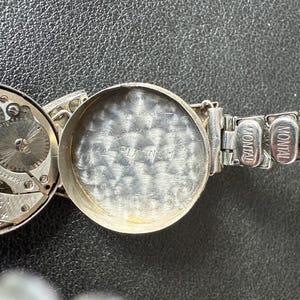 May include: Close-up of a silver-toned Montal watch with the back cover open, revealing the watch's inner workings and the inscription "PLATINUM". The watch band is also silver-toned.