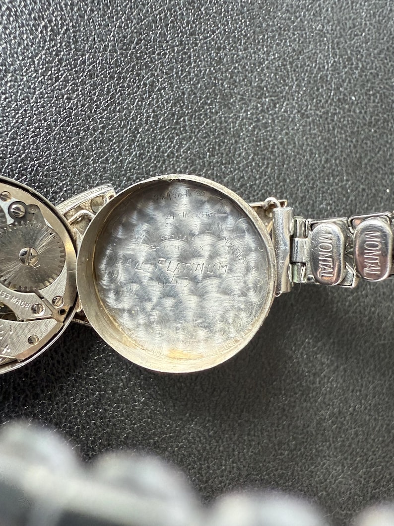 May include: Close-up of a silver-toned Montal watch with the back cover open, revealing the watch's inner workings and the inscription "PLATINUM". The watch band is also silver-toned.