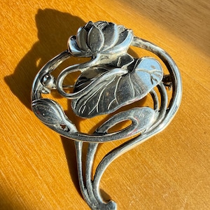 May include: A silver-toned brooch featuring a stylized lotus flower and leaves. The Art Nouveau-inspired design includes a central flower, a large leaf, and curved stems. The brooch is likely made of metal and has a pin closure on the back.