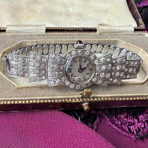 May include: An antique silver wristwatch with a bow-shaped face and band, encrusted with sparkling clear gemstones. The watch face has black numerals and hands. The watch is displayed in a vintage, beige lined box with a gold clasp.