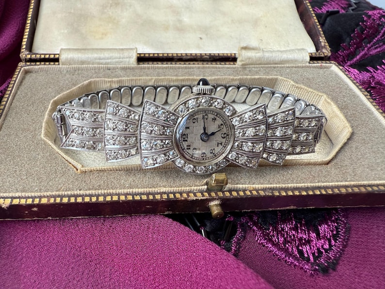 May include: An antique silver wristwatch with a bow-shaped face and band, encrusted with sparkling clear gemstones. The watch face has black numerals and hands. The watch is displayed in a vintage, beige lined box with a gold clasp.