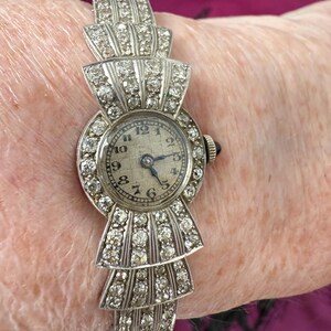 May include: An antique silver-toned wristwatch with a bow-shaped design. The watch face is round with a light-colored background and black numerals. The watch band is adorned with numerous small, sparkling stones. The watch is a vintage accessory.
