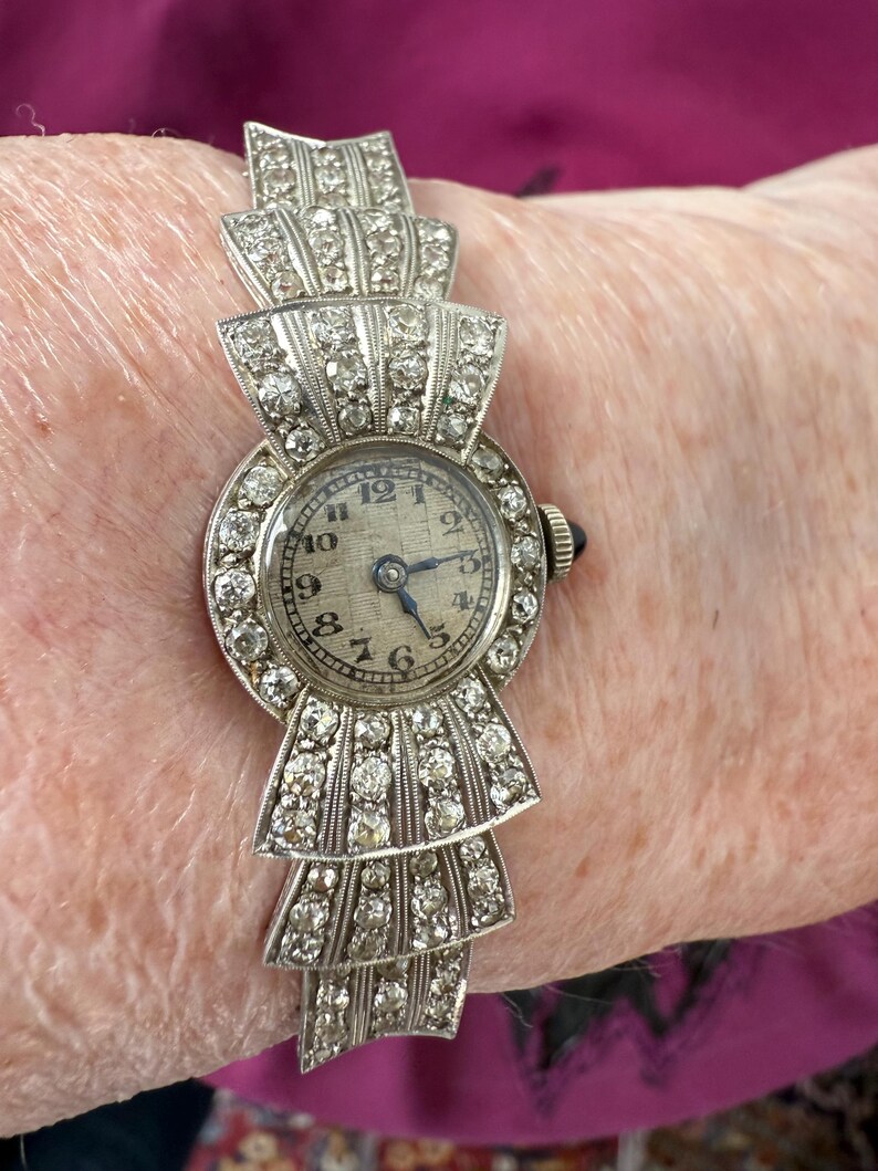 May include: An antique silver-toned wristwatch with a bow-shaped design. The watch face is round with a light-colored background and black numerals. The watch band is adorned with numerous small, sparkling stones. The watch is a vintage accessory.