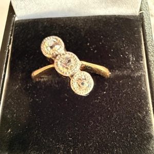 Könnte beinhalten: Ein goldener Ring mit drei runden, klaren Edelsteinen. Der Ring ist in einem goldenen Band gefasst und wird in einer schwarzen Samtbox präsentiert. Die Edelsteine sind gruppiert und bilden ein einzigartiges Design.