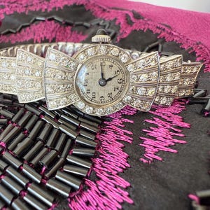 May include: A vintage silver-toned wristwatch with a bow-shaped face and band, encrusted with sparkling crystals. The watch face displays a classic analog clock with black hands and numerals. The watch is set against a dark background.