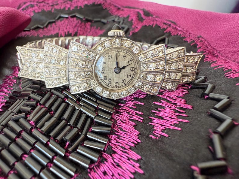 May include: A vintage silver-toned wristwatch with a bow-shaped face and band, encrusted with sparkling crystals. The watch face displays a classic analog clock with black hands and numerals. The watch is set against a dark background.