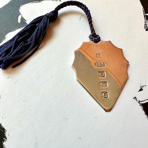 May include: A silver bookmark with a tassel. The bookmark is shaped like a shield and has the number 925 stamped on it.