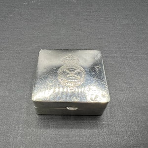 May include: A silver metal box with a hinged lid. The box has an embossed design on the top, featuring a crown and a winged emblem.