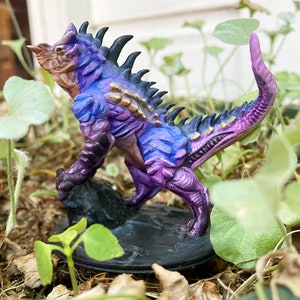 May include: Purple and gold painted miniature figurine of a reptilian creature with spikes and a long tail. The creature is standing on a black base.