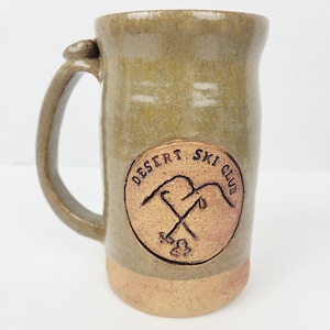 May include: A handmade ceramic mug with a large handle and a tan and olive green glaze. The mug features a circular emblem with the words "Desert Ski Club" and a crossed ski pole design.