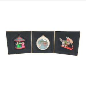 Peut inclure: Trois décorations de Noël dans des boîtes noires. L'une représente un carrousel avec des personnages, une autre une boule à neige avec un personnage, et la troisième un écureuil et un lapin sur une luge. Chaque ornement a un design et une palette de couleurs uniques.
