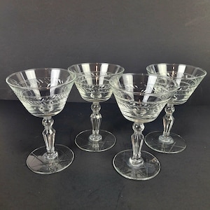 Puede incluir: Cuatro copas de champán transparentes con detalles grabados. Cada copa presenta un cuenco redondo, un tallo decorativo y una base circular. Las copas están dispuestas sobre una superficie oscura, mostrando su diseño vintage.
