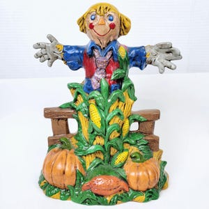 May include: A ceramic figurine of a scarecrow standing on a fence with corn stalks and pumpkins. The scarecrow is wearing a blue shirt, red vest, and a purple tie. The figurine is painted in bright colors.