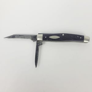May include: A folding pocket knife with a dark purple textured handle and silver metal bolsters. The knife features two metallic blades, one extended horizontally and one pointing downward, both showing signs of wear and oxidation.