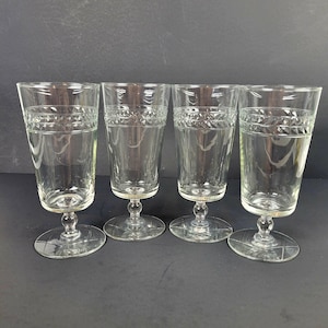 May include: Four clear, vintage water goblets. Each glass features a decorative band near the rim and a detailed stem. The glasses are displayed on a dark background, showcasing their elegant design. Perfect for a formal dinner setting.