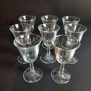 May include: A collection of eight clear glass cordial glasses. Each glass has a rounded bowl, a slender stem, and a circular base. The glasses are arranged on a dark surface, showcasing their transparent design and elegant shape.