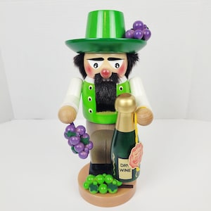 May include: A wooden nutcracker figurine of a man wearing a green hat and vest, holding a bunch of purple grapes and a green wine bottle. The figurine has a black beard and mustache, and is standing on a wooden base.