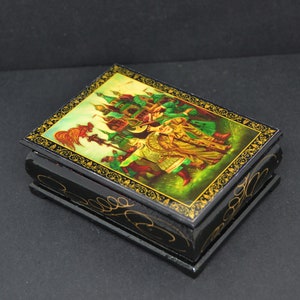 May include: A small wooden box with a hinged lid. The lid is decorated with a colorful painting of a scene from a Russian folk tale. The box is black with gold trim.