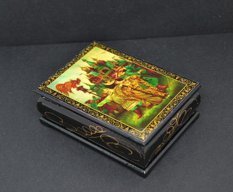 May include: A small wooden box with a hinged lid. The lid is decorated with a colorful painting of a scene from a Russian folk tale. The box is black with gold trim.