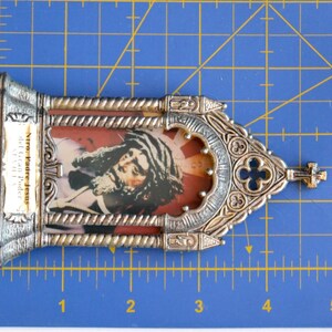 May include: A silver-toned religious icon featuring an image of Jesus with long hair and a beard, set against a red background. The frame has ornate detailing and a small cross at the top. The text "Ntro. Padre Jesus del Gran Poder" is visible.