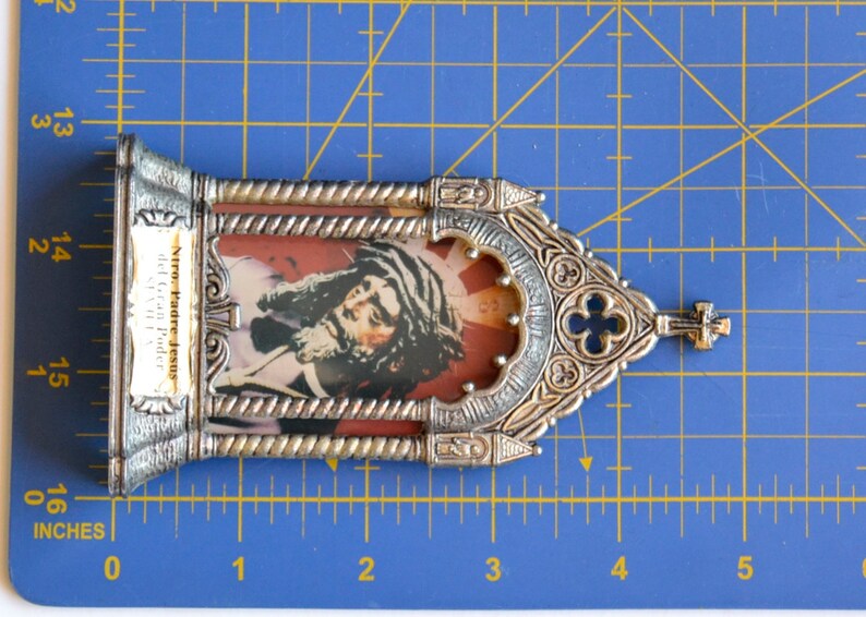 May include: A silver-toned religious icon featuring an image of Jesus with long hair and a beard, set against a red background. The frame has ornate detailing and a small cross at the top. The text "Ntro. Padre Jesus del Gran Poder" is visible.