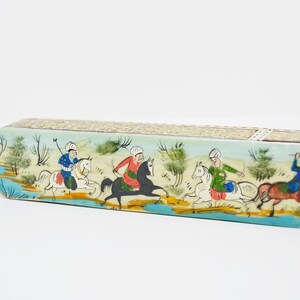 Puede incluir: Una caja larga y ovalada con una escena pintada de cuatro personas montando a caballo. La caja est&aacute; decorada con un fondo azul claro, caballos blancos y ropa colorida en los jinetes. La caja est&aacute; hecha de madera y tiene una superficie texturizada.