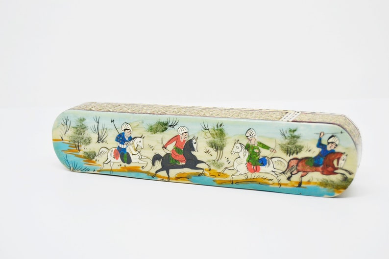 Puede incluir: Una caja larga y ovalada con una escena pintada de cuatro personas montando a caballo. La caja est&aacute; decorada con un fondo azul claro, caballos blancos y ropa colorida en los jinetes. La caja est&aacute; hecha de madera y tiene una superficie texturizada.