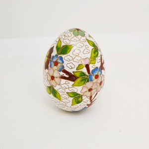 May include: A white egg-shaped trinket box with a floral design. The box is decorated with pink, blue, green, and gold enamel.