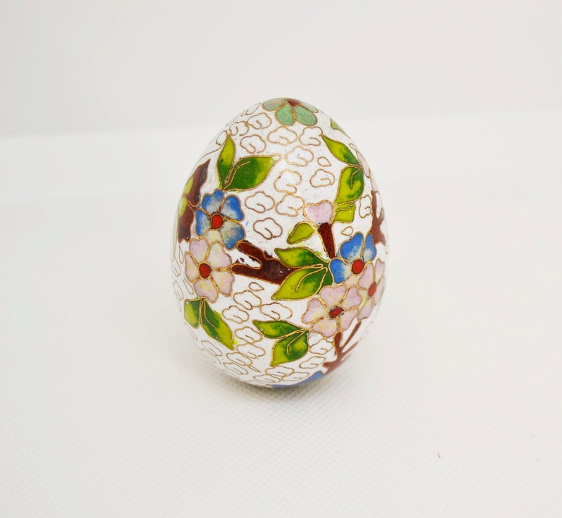 May include: A white egg-shaped trinket box with a floral design. The box is decorated with pink, blue, green, and gold enamel.