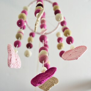May include: A pink and beige baby mobile with crocheted hearts and felt balls hanging from a circular metal frame.