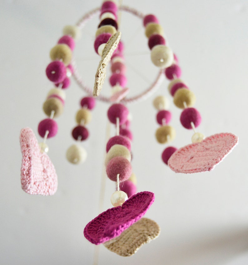 May include: A pink and beige baby mobile with crocheted hearts and felt balls hanging from a circular metal frame.