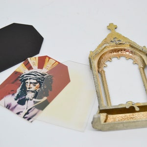 May include: An antique gold-colored metal frame with a cross on top, next to a black tag and two tags with a religious image. The image depicts a man with a crown of thorns and a red background.