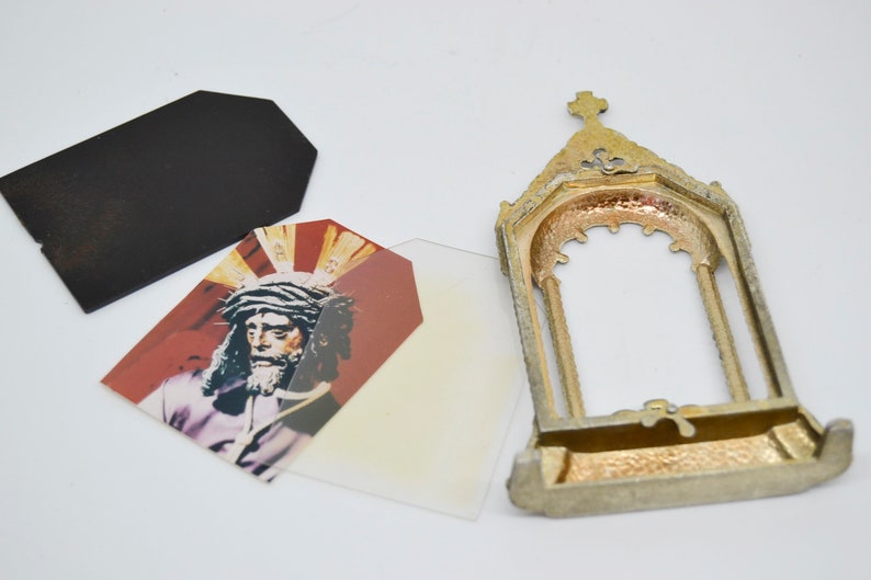 May include: An antique gold-colored metal frame with a cross on top, next to a black tag and two tags with a religious image. The image depicts a man with a crown of thorns and a red background.