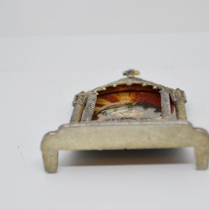 May include: A small, ornate silver-toned religious shrine with a detailed arched top and a small cross. The shrine features a colorful image within, set against a red backdrop. The base has a rectangular shape.