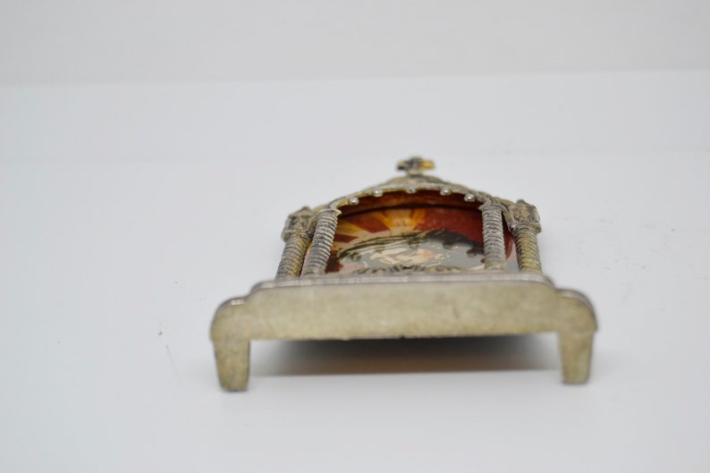 May include: A small, ornate silver-toned religious shrine with a detailed arched top and a small cross. The shrine features a colorful image within, set against a red backdrop. The base has a rectangular shape.