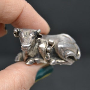 May include: A silver cow figurine, lying down with its head turned to the side. The figurine is detailed with a textured surface and has a small chain around its neck.