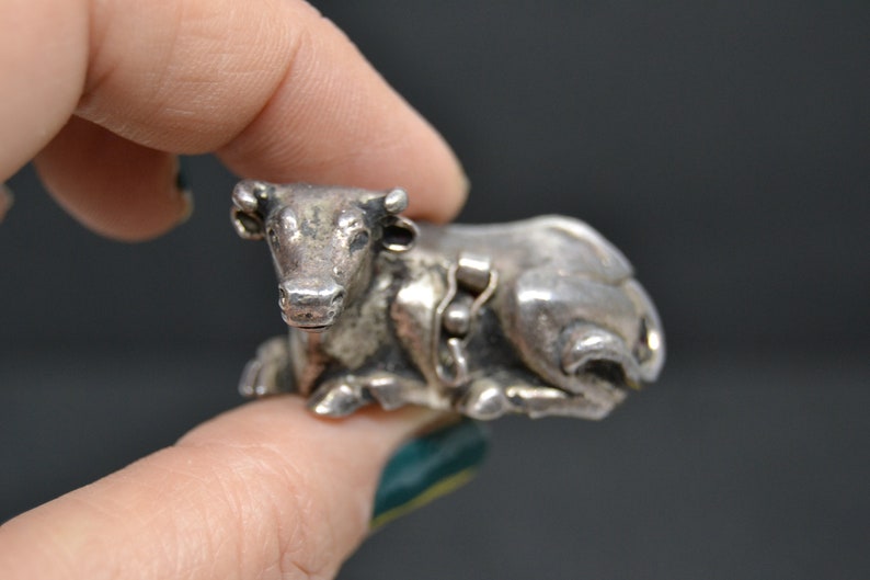 May include: A silver cow figurine, lying down with its head turned to the side. The figurine is detailed with a textured surface and has a small chain around its neck.