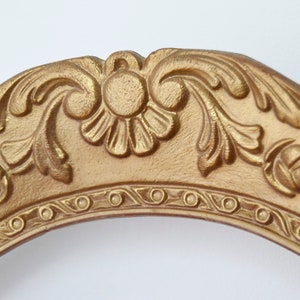 May include: An ornate picture frame in a gold colour with a decorative, raised, floral design. The frame is oval shaped and has a smooth, polished finish.