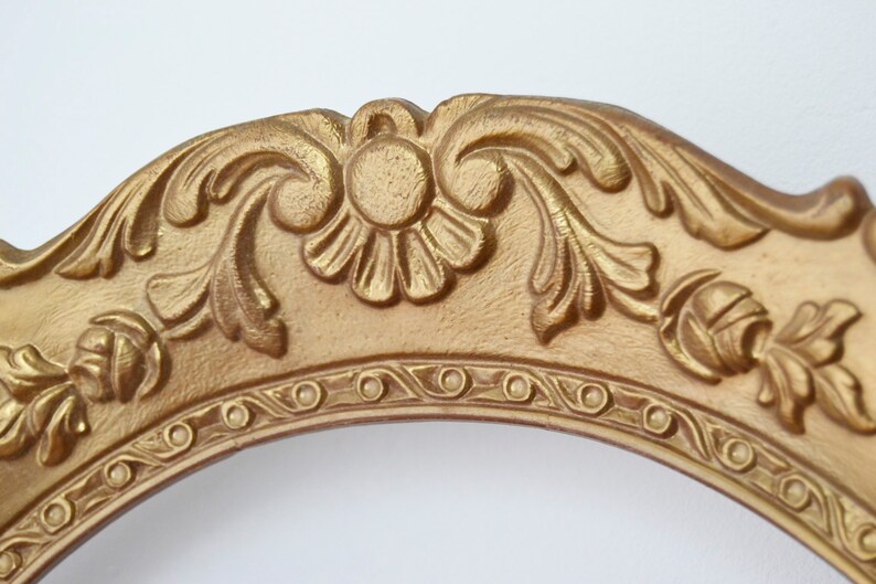 May include: An ornate picture frame in a gold colour with a decorative, raised, floral design. The frame is oval shaped and has a smooth, polished finish.