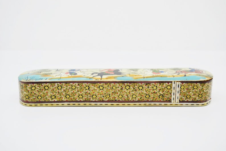 Puede incluir: Una caja larga y rectangular con un dise&ntilde;o colorido e intrincado. La caja est&aacute; hecha de madera y tiene una tapa con bisagras. La parte superior de la caja presenta una escena de personas montando a caballo, con un fondo azul y blanco. La parte inferior de la caja tiene un patr&oacute;n repetitivo de flores y formas geom&eacute;tricas.