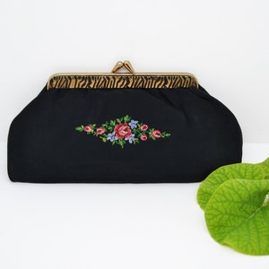 Puede incluir: Un bolso de mano negro de tela con un cierre de metal dorado y un diseño de bordado floral. El bordado presenta rosas rosas y rojas con hojas verdes y flores azules.