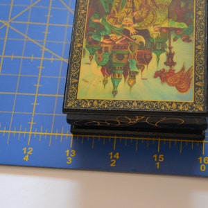 May include: A small, black wooden box with a hinged lid. The lid is decorated with a colorful painting of a scene with buildings, people, and a bird. The box is about 4 inches wide and 3 inches tall.